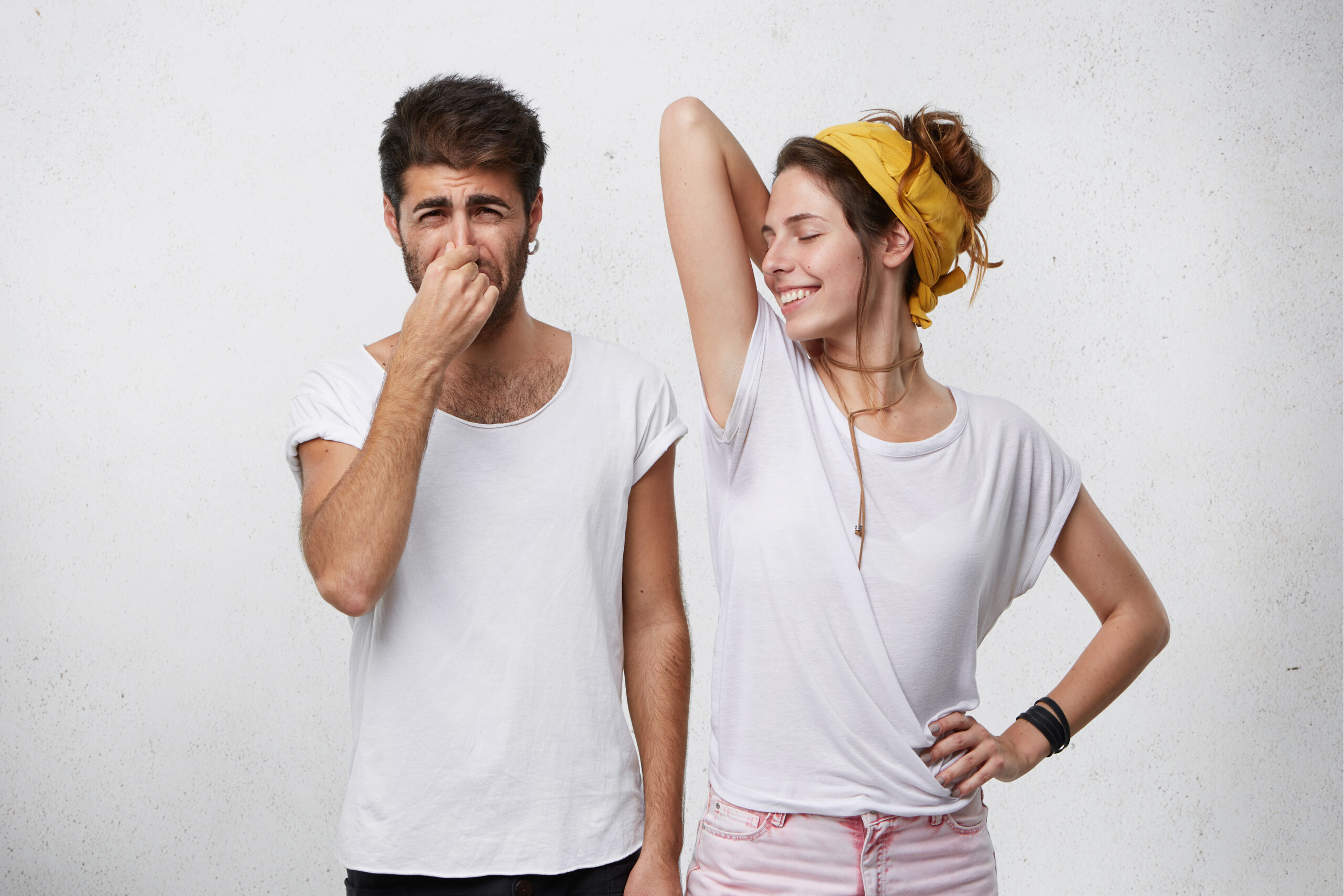 Problems with body odor. Disgusted male pinching his nose feeling bad smell or stink coming out from attractive smiling girl, who is raising her arm, showing wet t-shirt because of armpit sweat