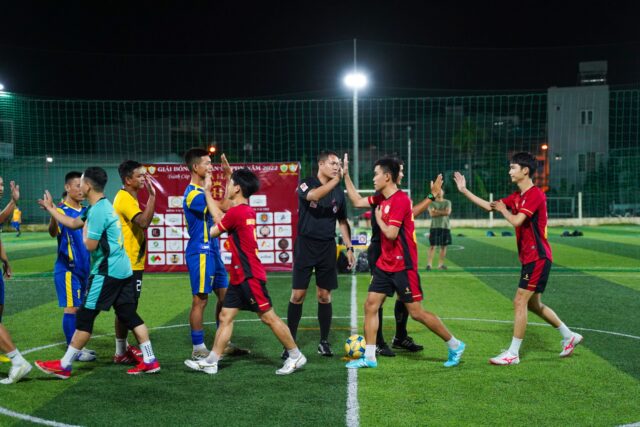 Arbitru de fotbal pe gazonul unui stadion, în timpul unui meci, lângă jucătorii celor două echipe