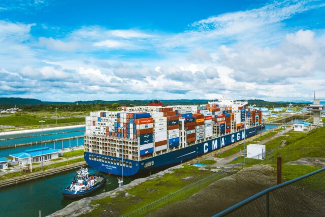 Navă comercială încărcată cu containere care traversează un canal maritim îngust între două maluri